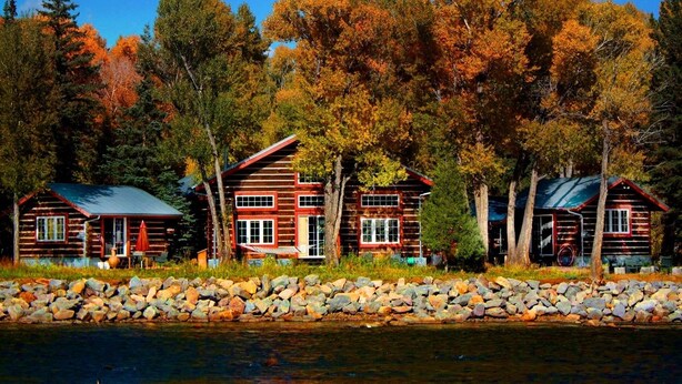 A log cabin resort with multiple cabins by a lake, surrounded by autumn foliage. - Riverside Meadows Cabins (South Fork)