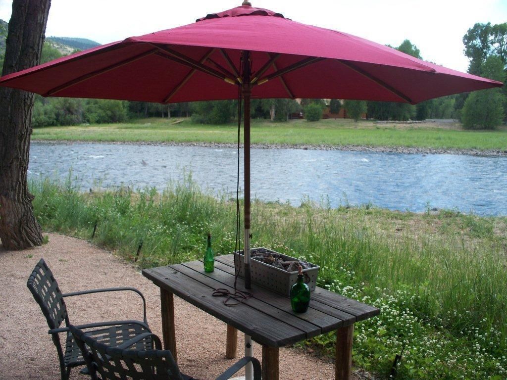 Un coin pique-nique aménagé avec une table et des chaises près d’une rivière, un parasol rouge et une bouteille de vin.