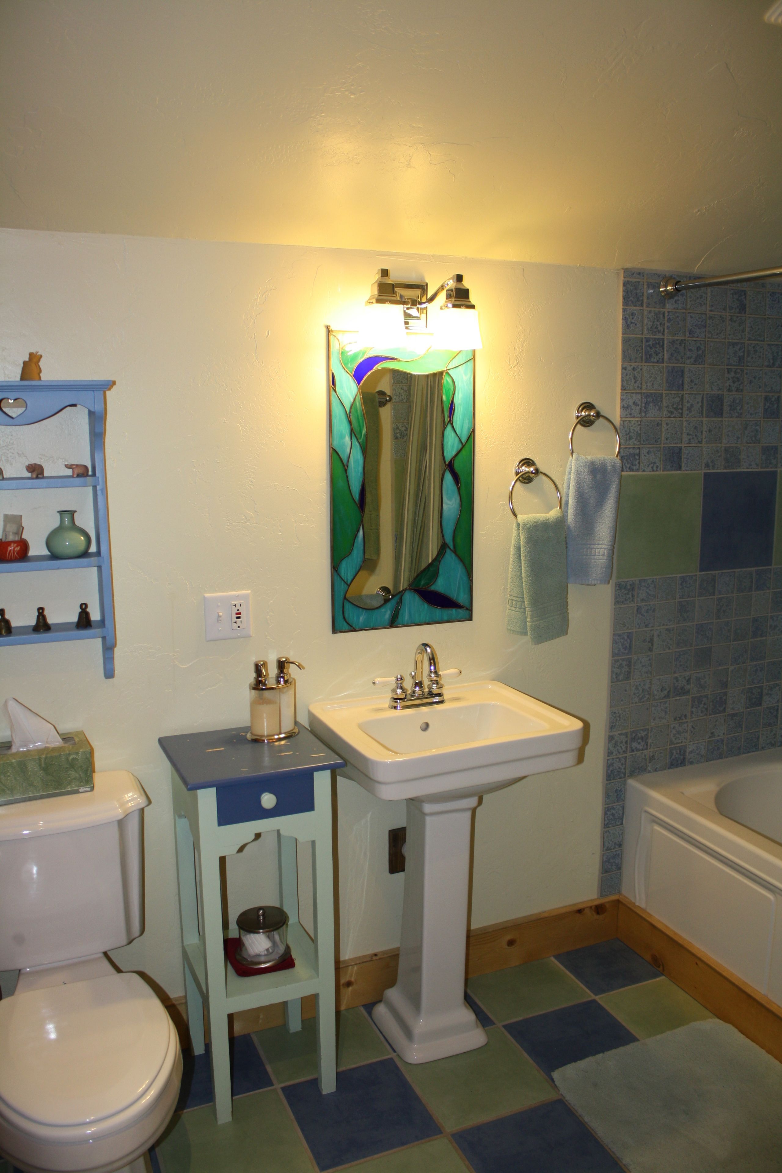 Un baño con lavamanos blanco, inodoro, repisa azul con artículos de aseo, espejo de vitral y zona de ducha con azulejos azules.