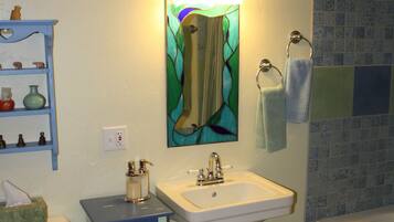 Un baño con lavamanos blanco, inodoro, repisa azul con artículos de aseo, espejo de vitral y zona de ducha con azulejos azules.