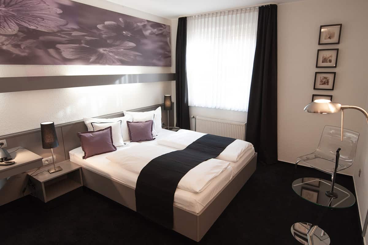 A hotel room with a large bed, a nightstand, a desk, a chair, a lamp, a window with curtains, and a wall with framed pictures.