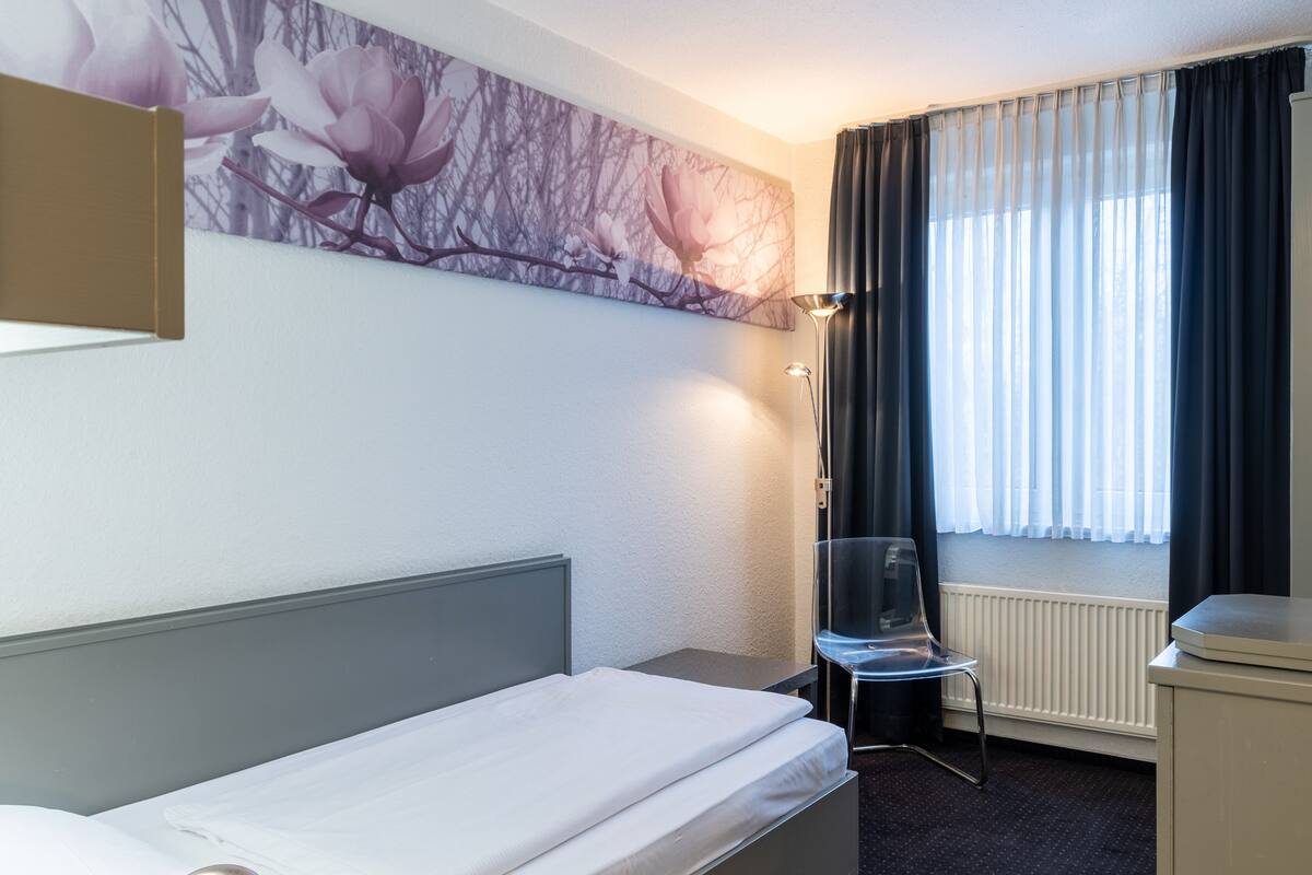 A hotel room with a bed, a chair, a desk, a radiator, and a window with curtains.