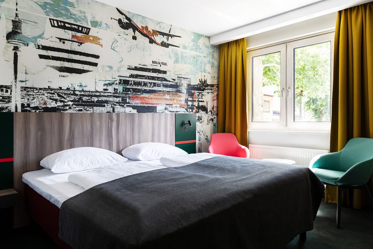 A hotel room with a large bed, a red chair, a teal chair, and a mural of Berlin's skyline.