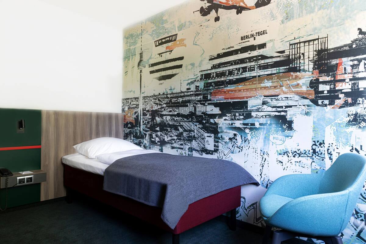 A hotel room with a bed, a chair, a wall-mounted phone, and a large cityscape mural.