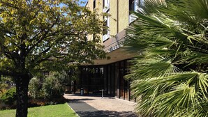 Un bâtiment moderne à la façade vitrée, entouré d’un jardin bien entretenu où l’on trouve un palmier et un grand arbre aux feuilles vertes.