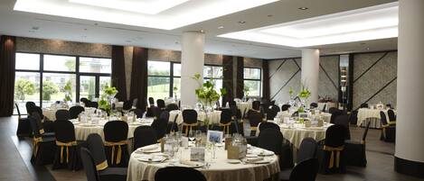 A large banquet hall with round tables, black chairs with gold sashes, and large windows offering a view of the outdoors.