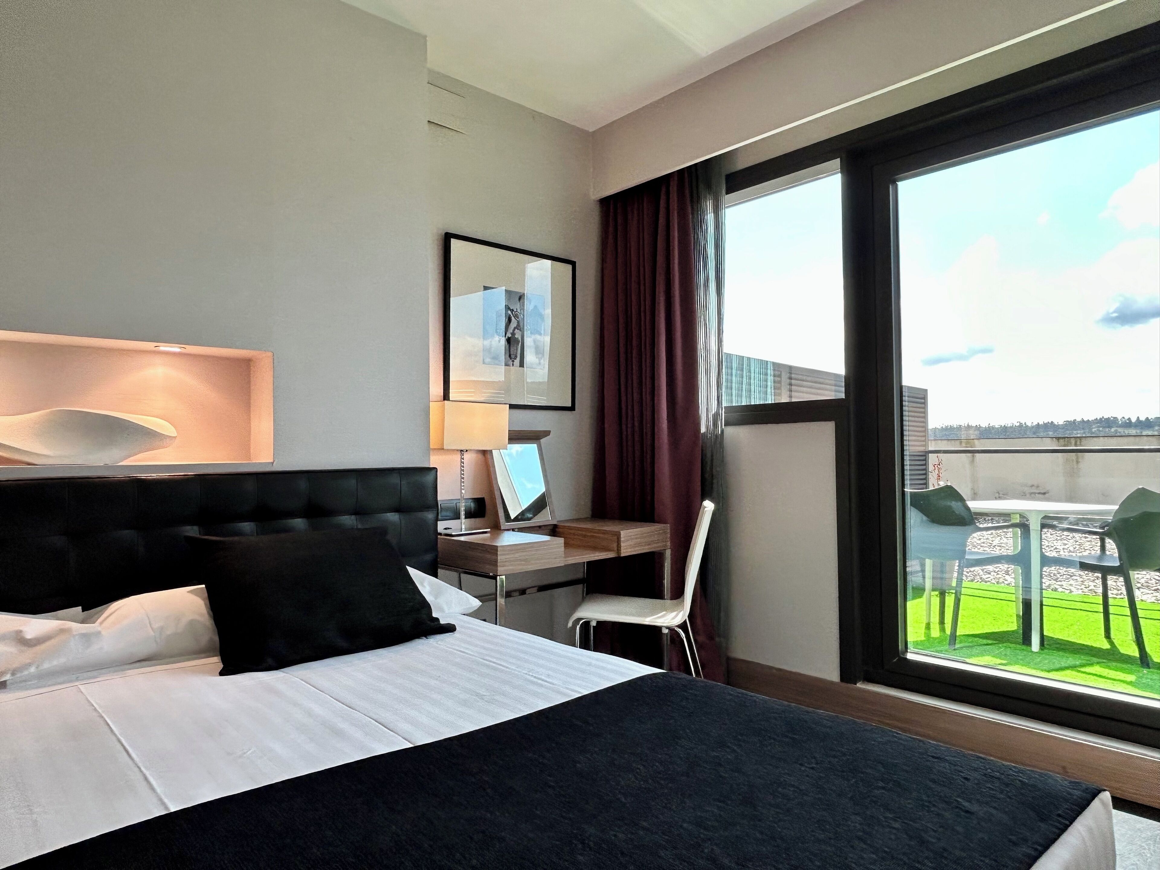 A hotel room with a large bed, a desk with a computer, a chair, and a view of a balcony with outdoor seating.
