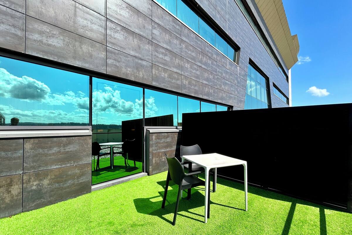Ein moderner Außenbereich zum Sitzen mit Tisch und Stühlen, eine Glaswand mit Blick auf Himmel und Wolken sowie ein Belag aus synthetischem Grünrasen.