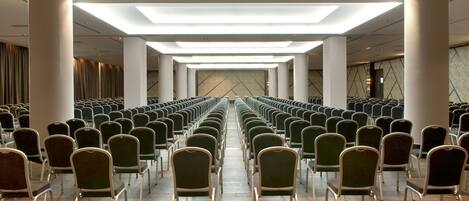Un salón de conferencias con filas de sillas, un techo alto con tragaluces y un papel tapiz en el fondo.