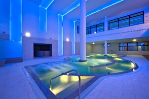 An indoor swimming pool with a central water feature, surrounded by a tiled floor and white pillars.