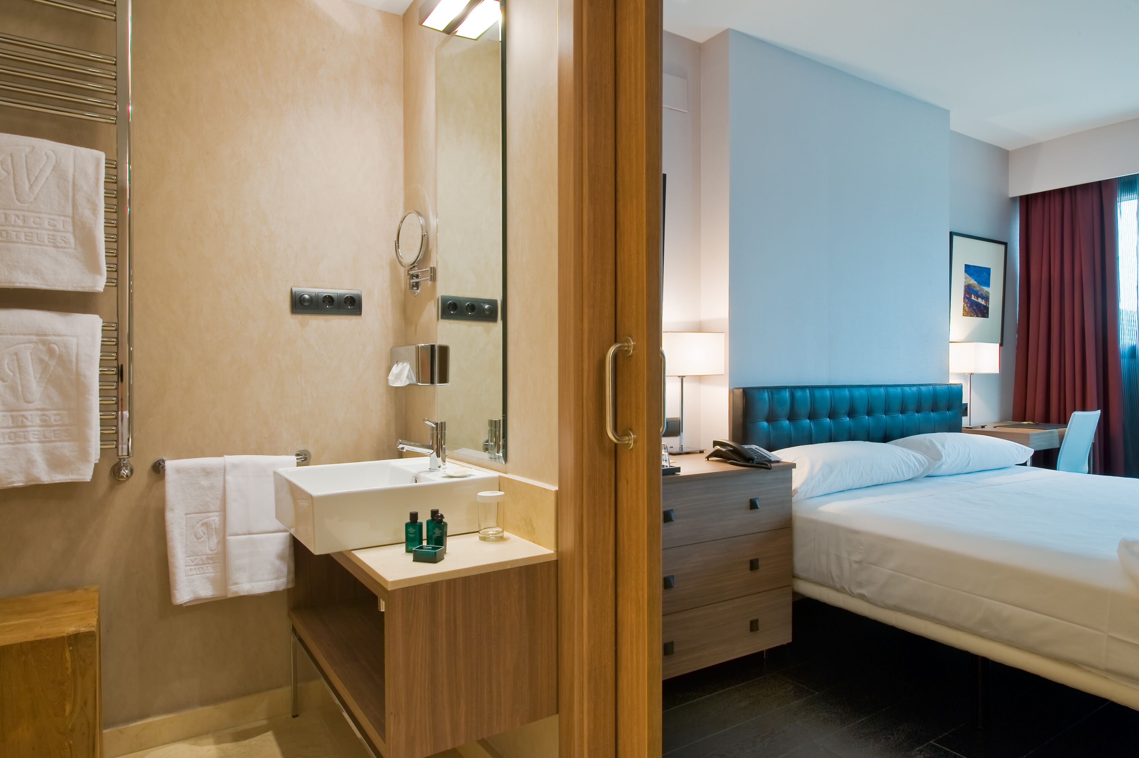 A hotel room with a wooden vanity, a large mirror, a bed with a blue headboard, and a bedside table with a lamp.