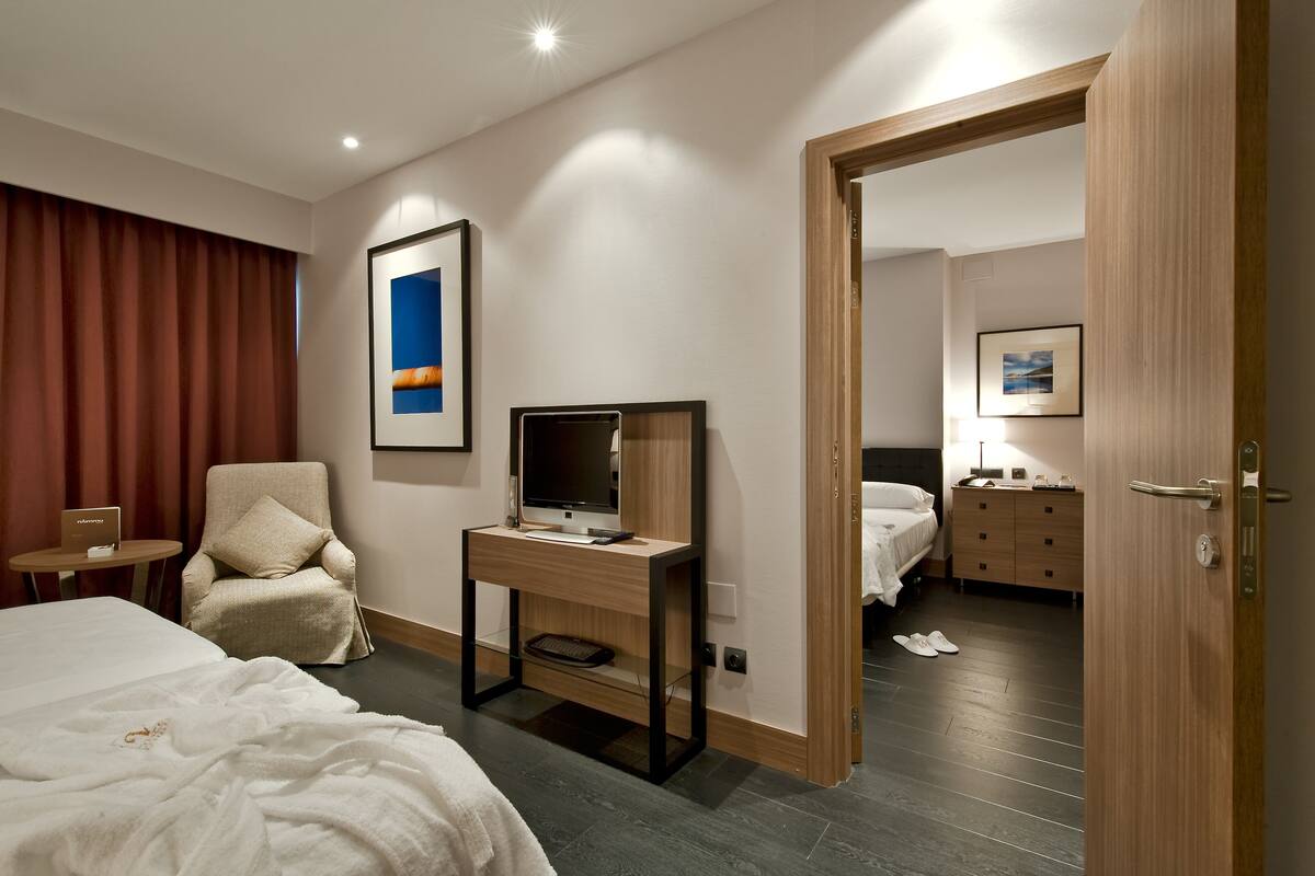 Ein Hotelzimmer mit einem Bett, einem Schreibtisch mit Computer, einem Sessel, einem Fernseher und einer Tür zu einem weiteren Raum.