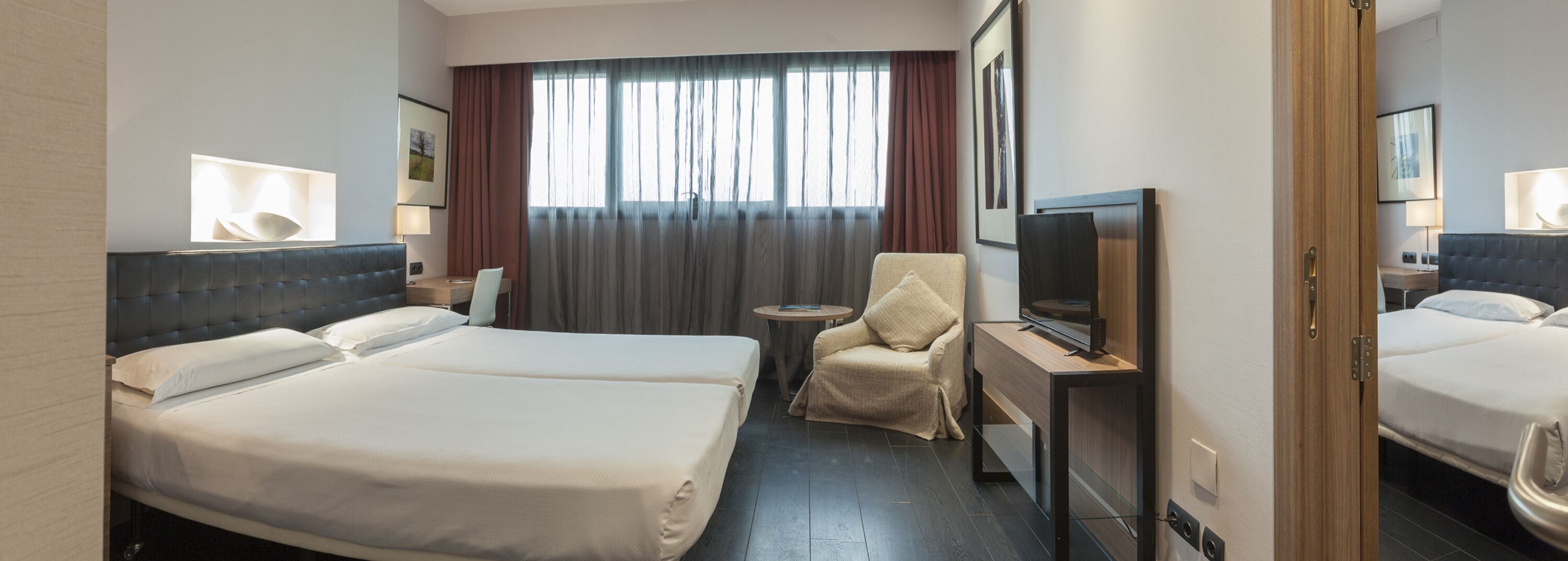 A hotel room with two beds, a TV, a chair, and a window with curtains.