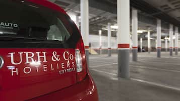 A red car with "URH & Co Hotelers" and "AUTO FIGUERES" on the rear window, parked in a parking garage with concrete pillars and a ceiling.