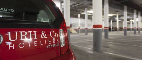 En röd bil med texten ”URH & Co Hotelers” och ”AUTO FIGUERES” på bakrutan, parkerad i ett parkeringsgarage med betongpelare och ett tak.