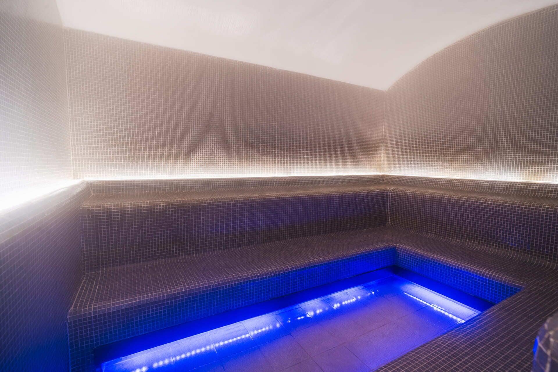 A spa room with a tiled floor and walls, a seating area with a blue-lit water feature, and a ceiling with a textured pattern.