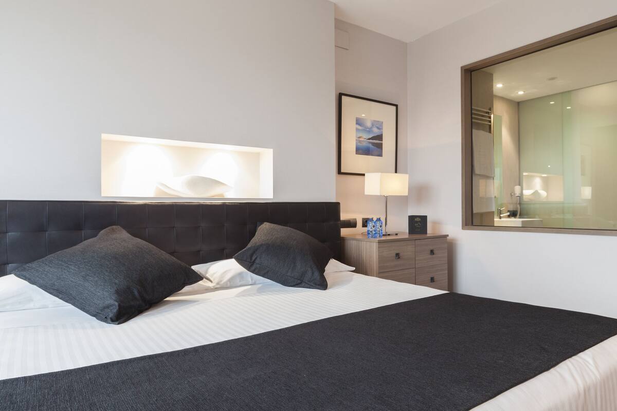Ein modernes Hotelzimmer mit einem großen Bett, einem Nachttisch mit Lampe, einem gerahmten Bild an der Wand und einem großen Fenster.