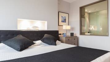A modern hotel room with a large bed, a bedside table with a lamp, a framed picture on the wall, and a large window.