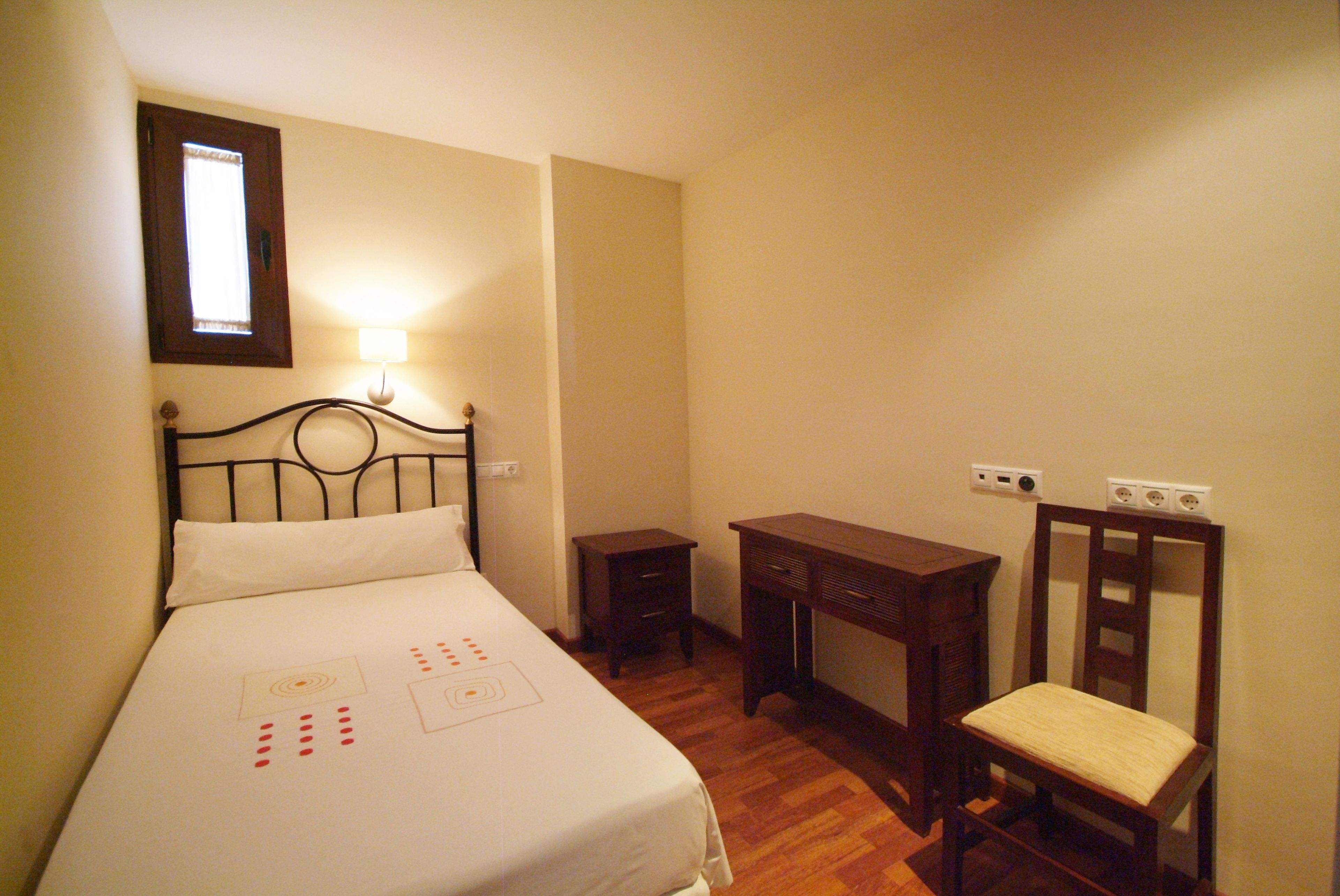 A bedroom with a bed, bedside table, desk, chair, and a wall-mounted lamp.