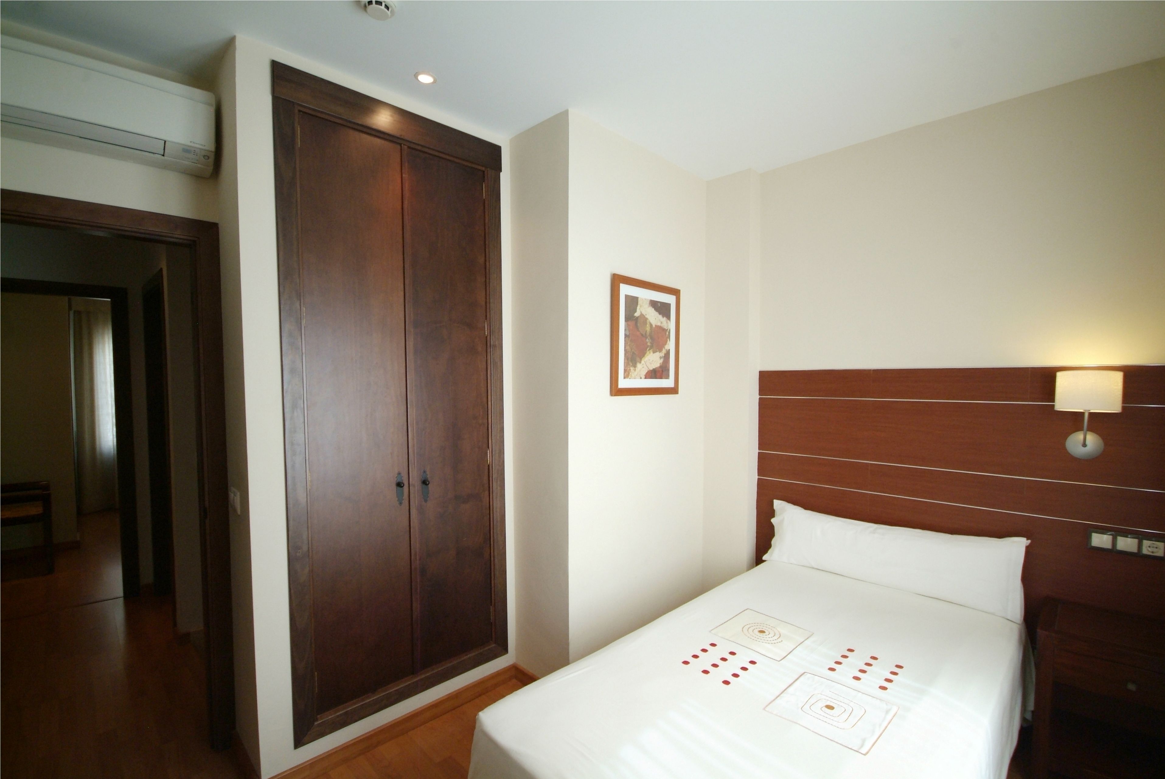 A hotel room with a large wooden wardrobe, a neatly made bed with a white bedspread, and a wall-mounted light fixture.