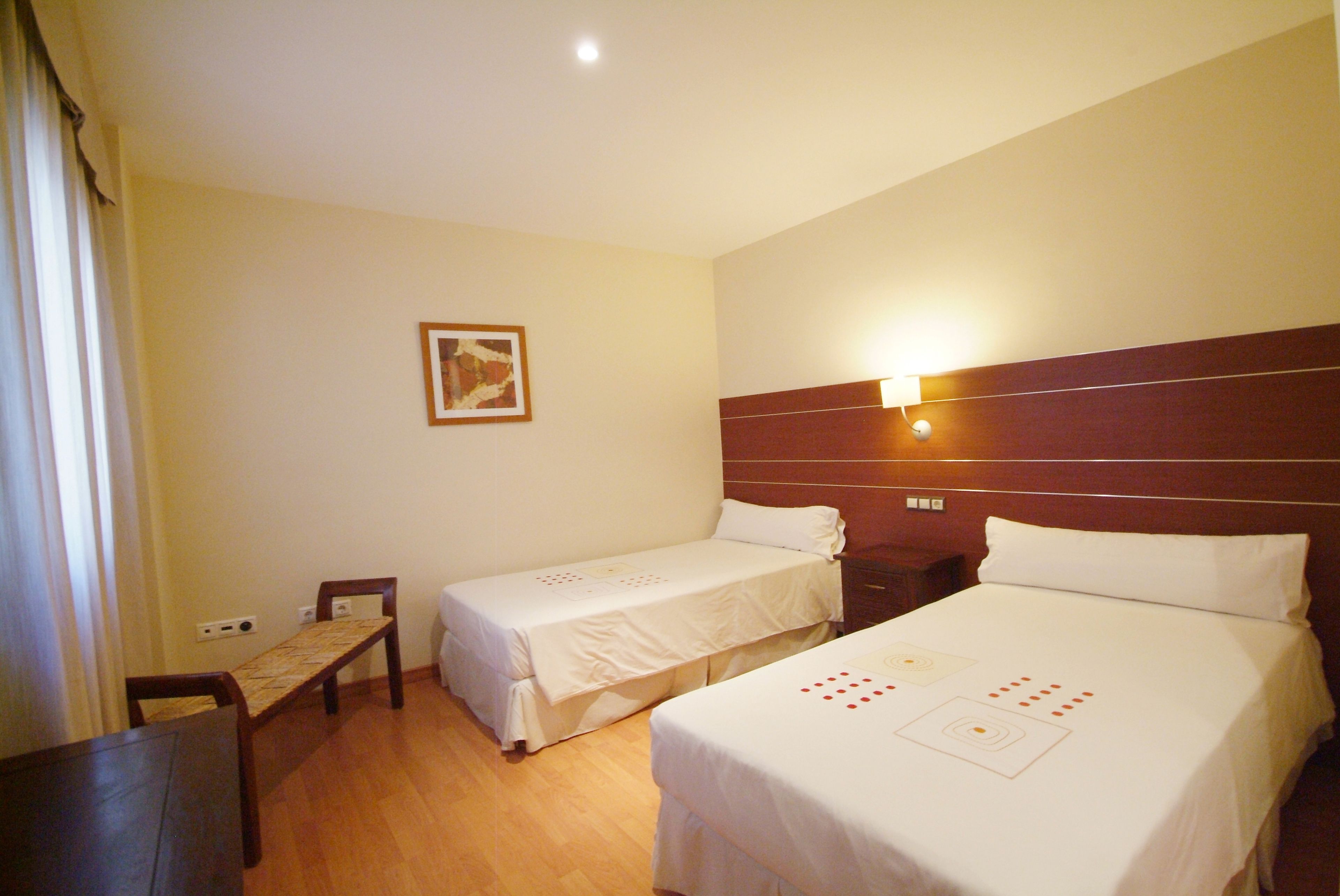 Una habitación de hotel con dos camas individuales, un banco de madera, una mesita pequeña y una lámpara fijada a la pared.