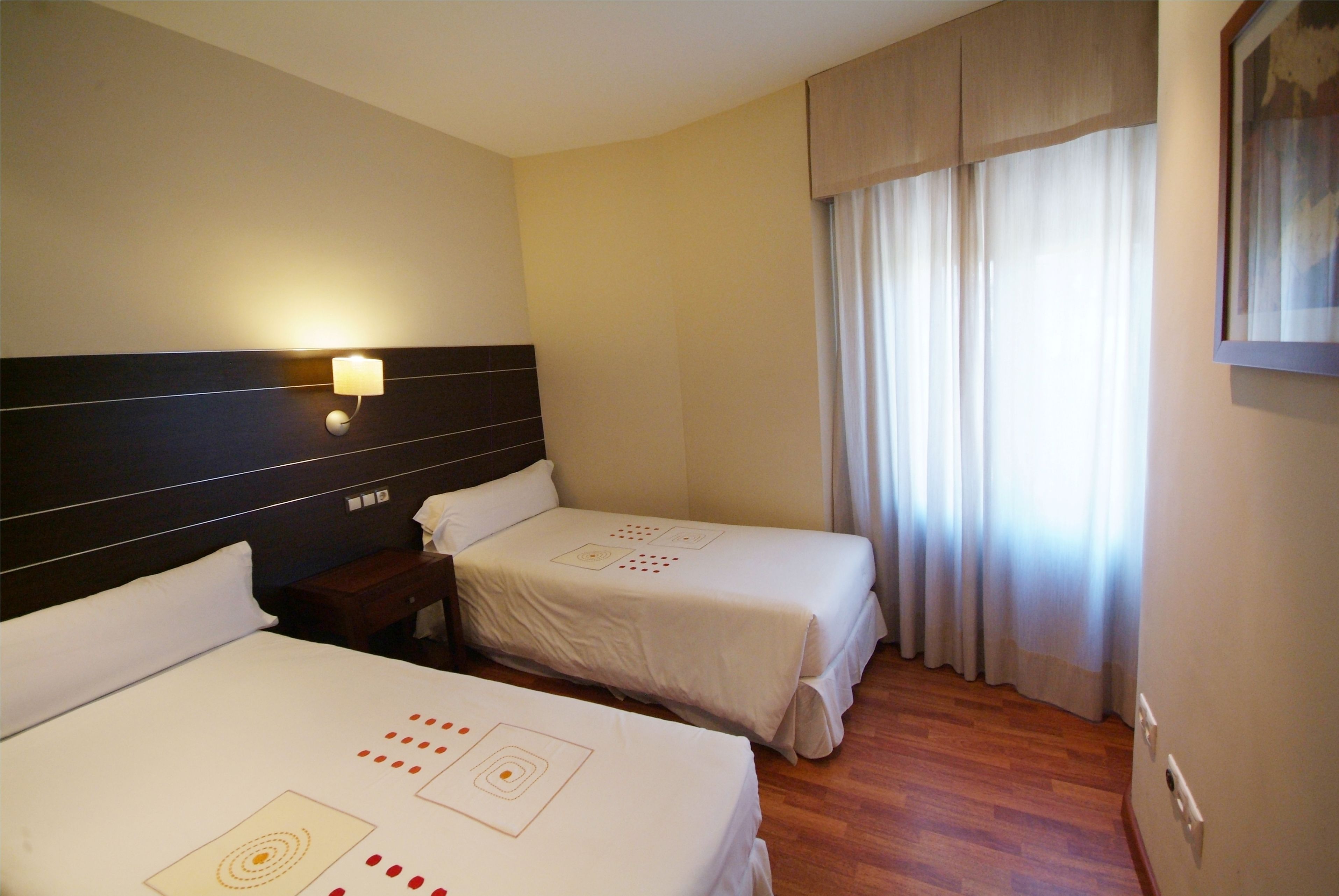 Une chambre d’hôtel avec deux lits, une table de chevet, une lampe fixée au mur et une fenêtre avec des rideaux.