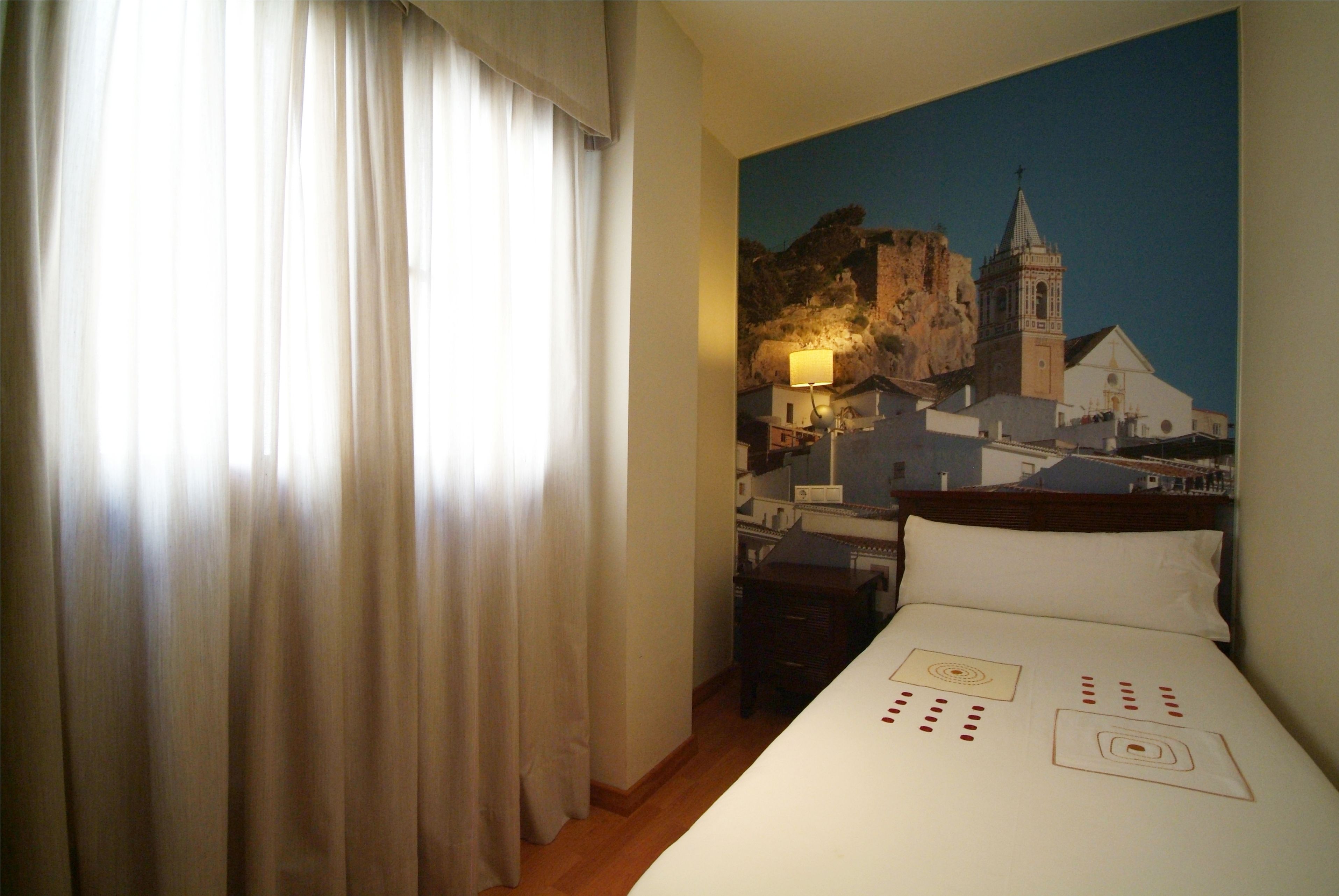 A hotel room with a bed, a nightstand, a lamp, and a painting of a coastal town.