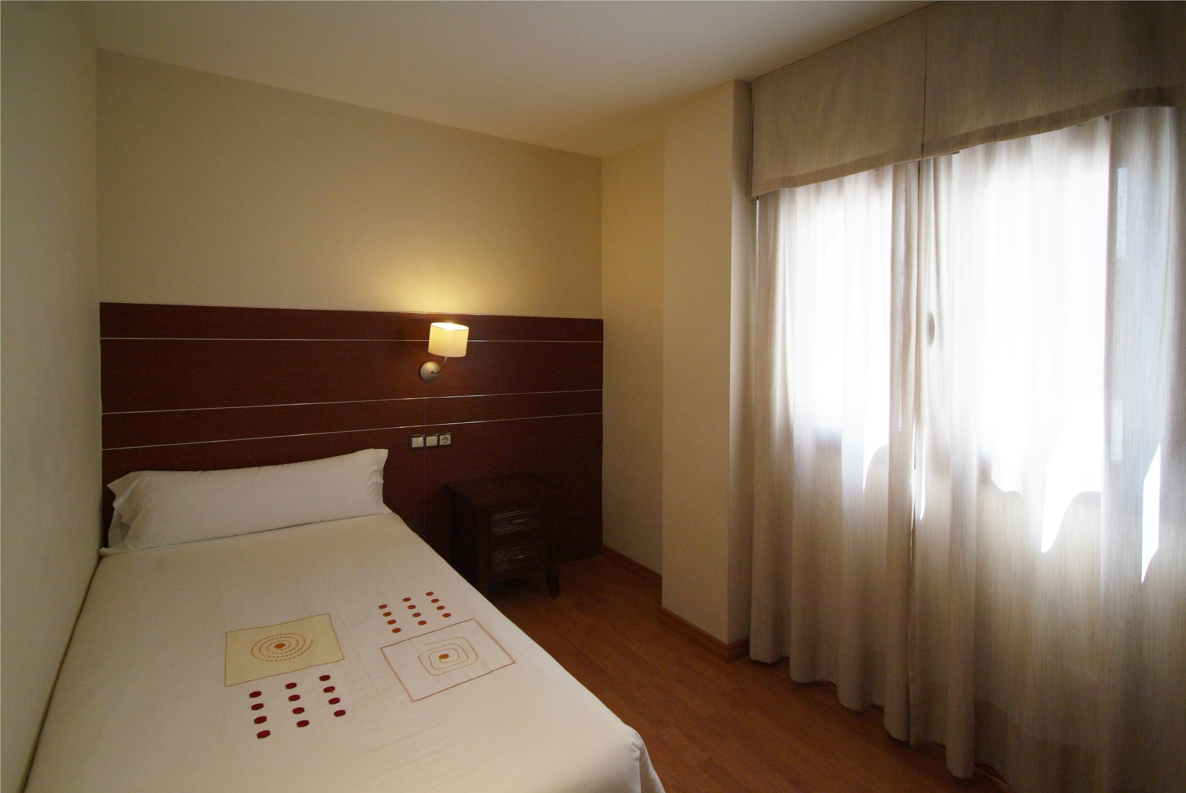 Una habitación de hotel con una cama bien hecha, una mesita de noche, una lámpara y una ventana con cortinas.