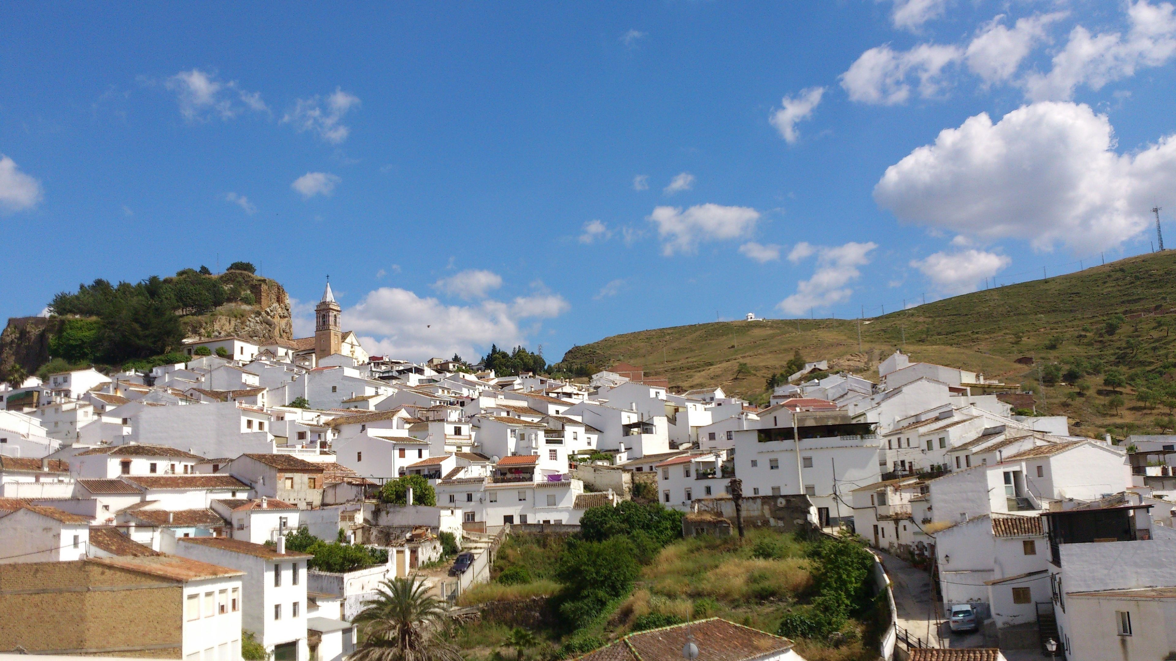 A densely packed town with white buildings, a prominent church with a bell tower, and a hill in the background.