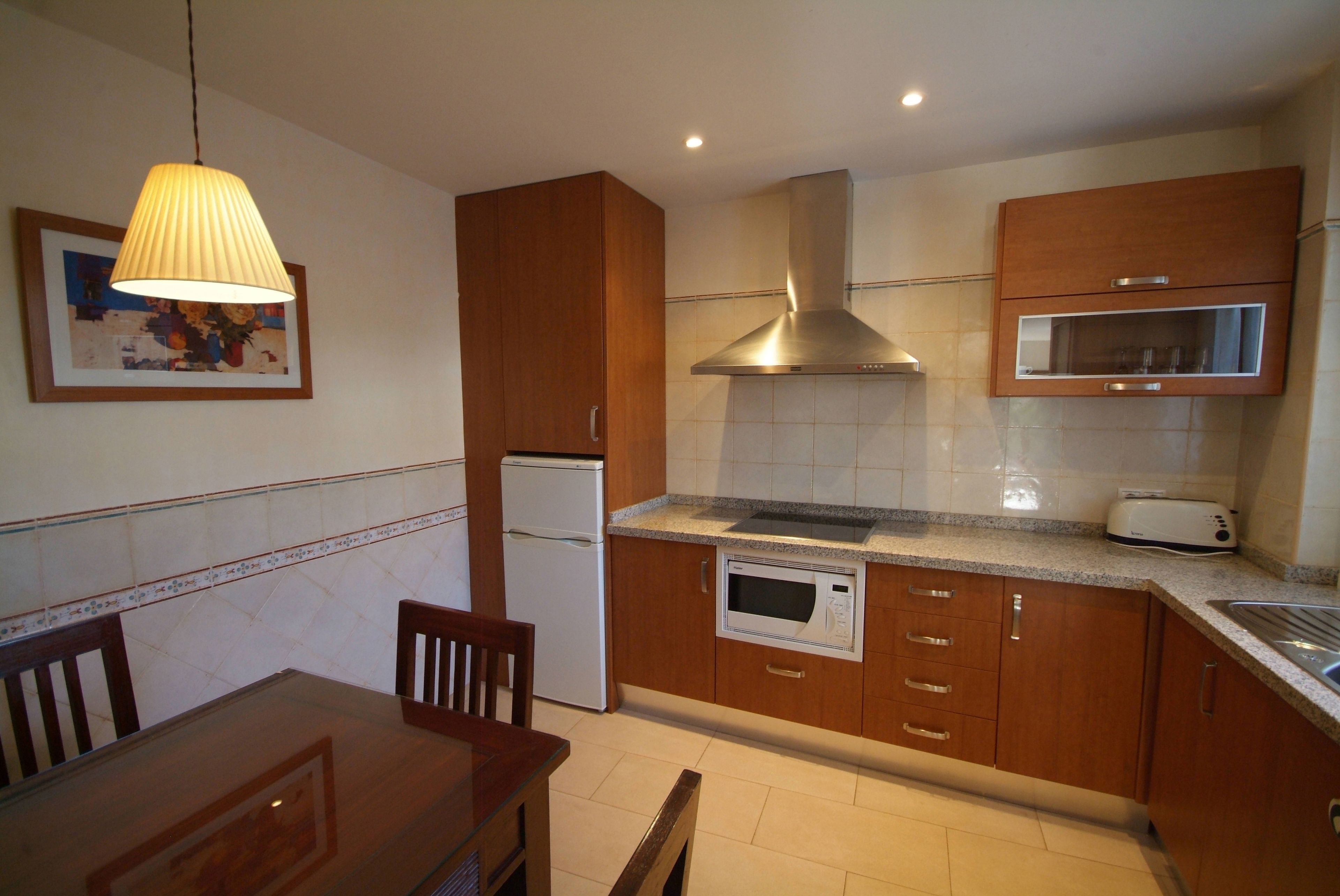 A compact kitchen with wooden cabinets, a built-in microwave, a stainless steel range hood, and a dining area with a table and chairs.