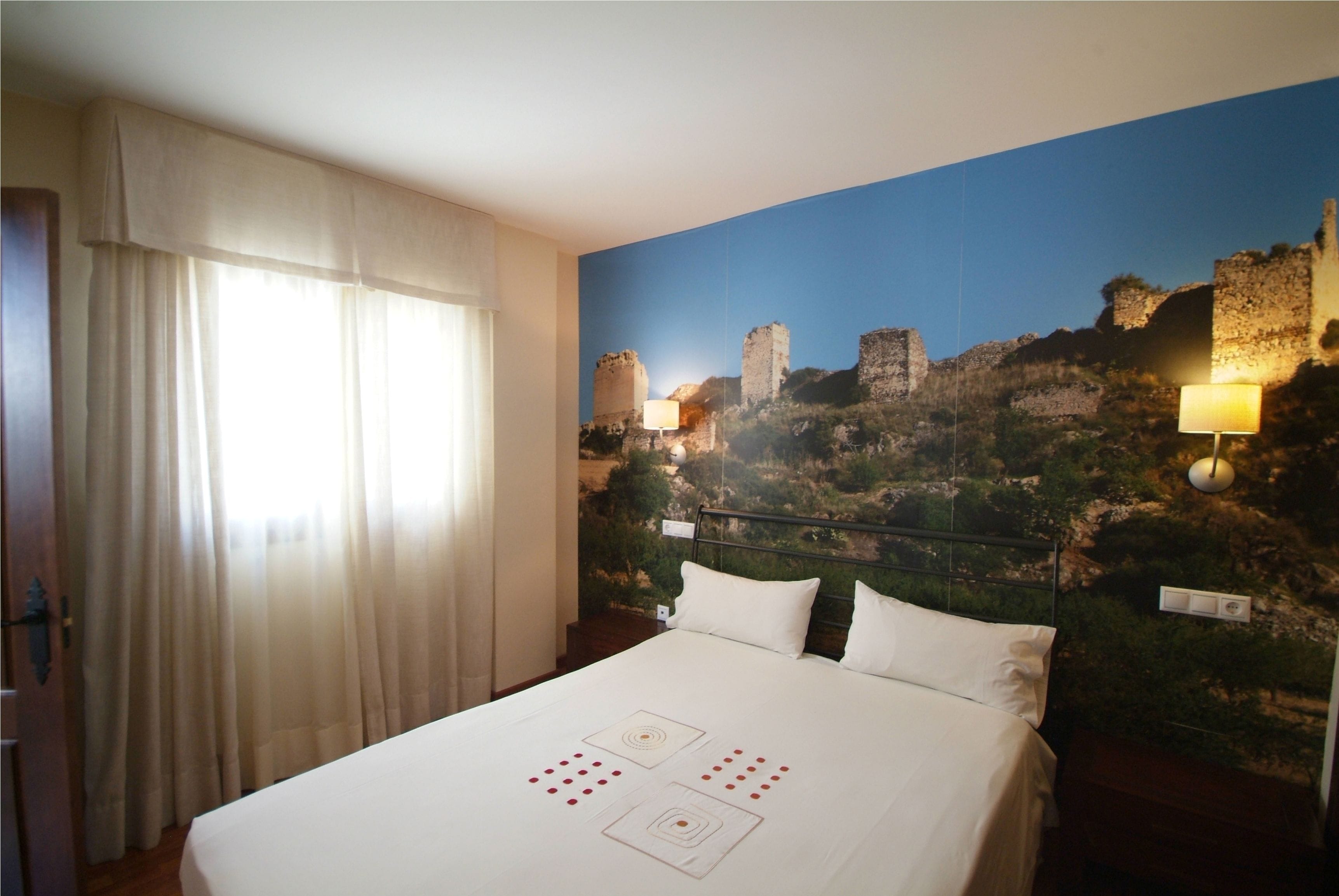 Habitación de hotel con cama, cortinas blancas, un mural de un sitio histórico y dos lámparas de pared.