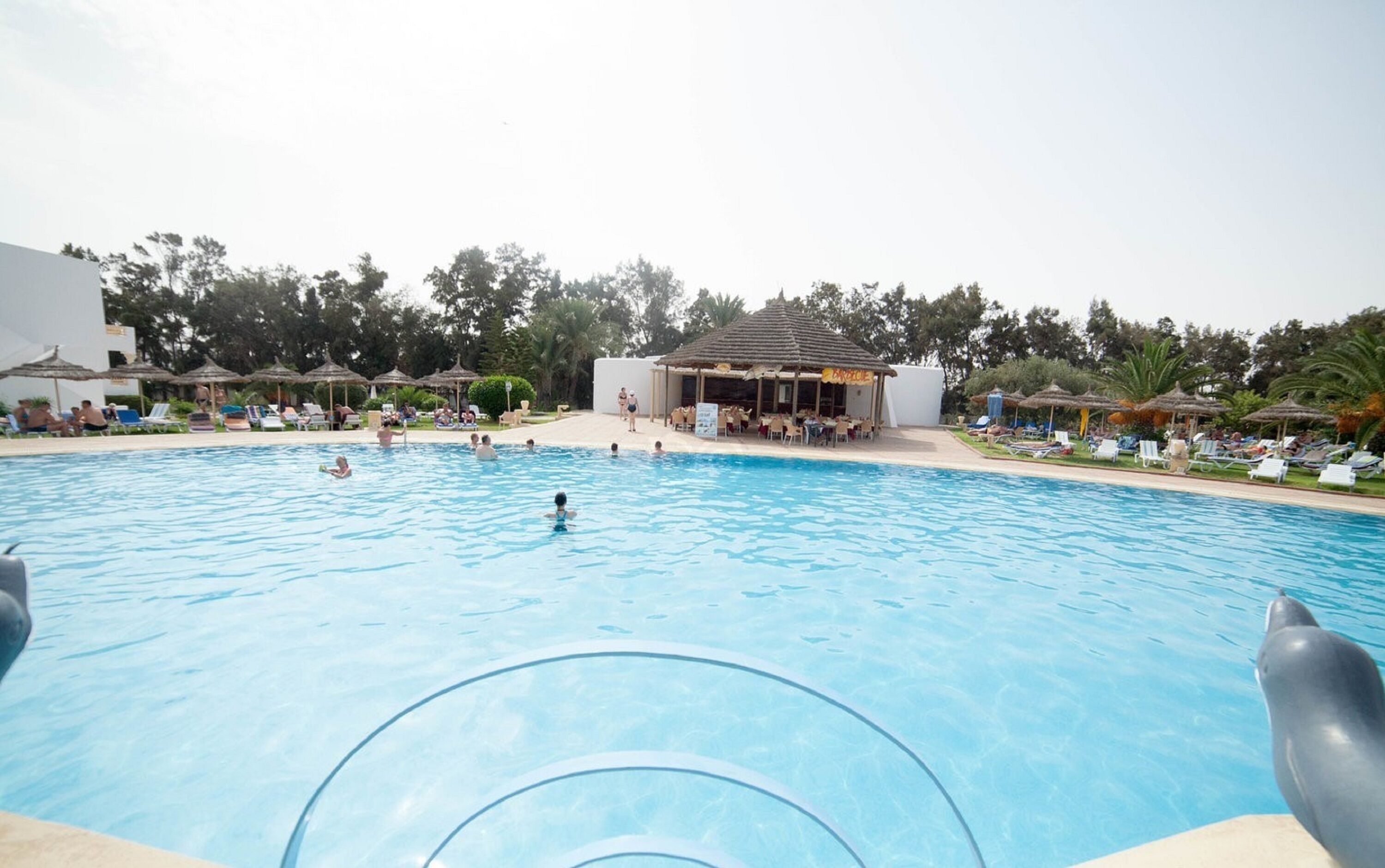 Piscina con sillones de playa y sombrillas, rodeada de árboles y un edificio con techo de paja.