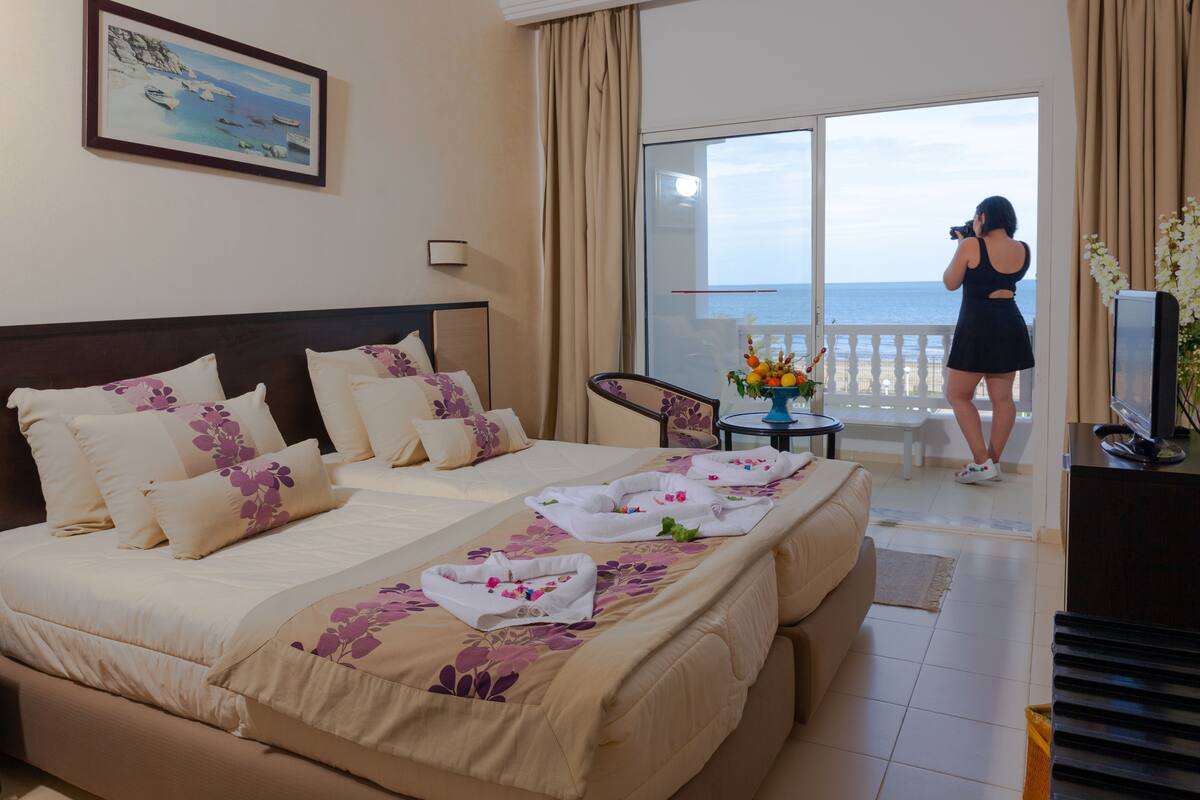 A hotel room with a bed, a balcony with a view of the sea, and a television on a stand.