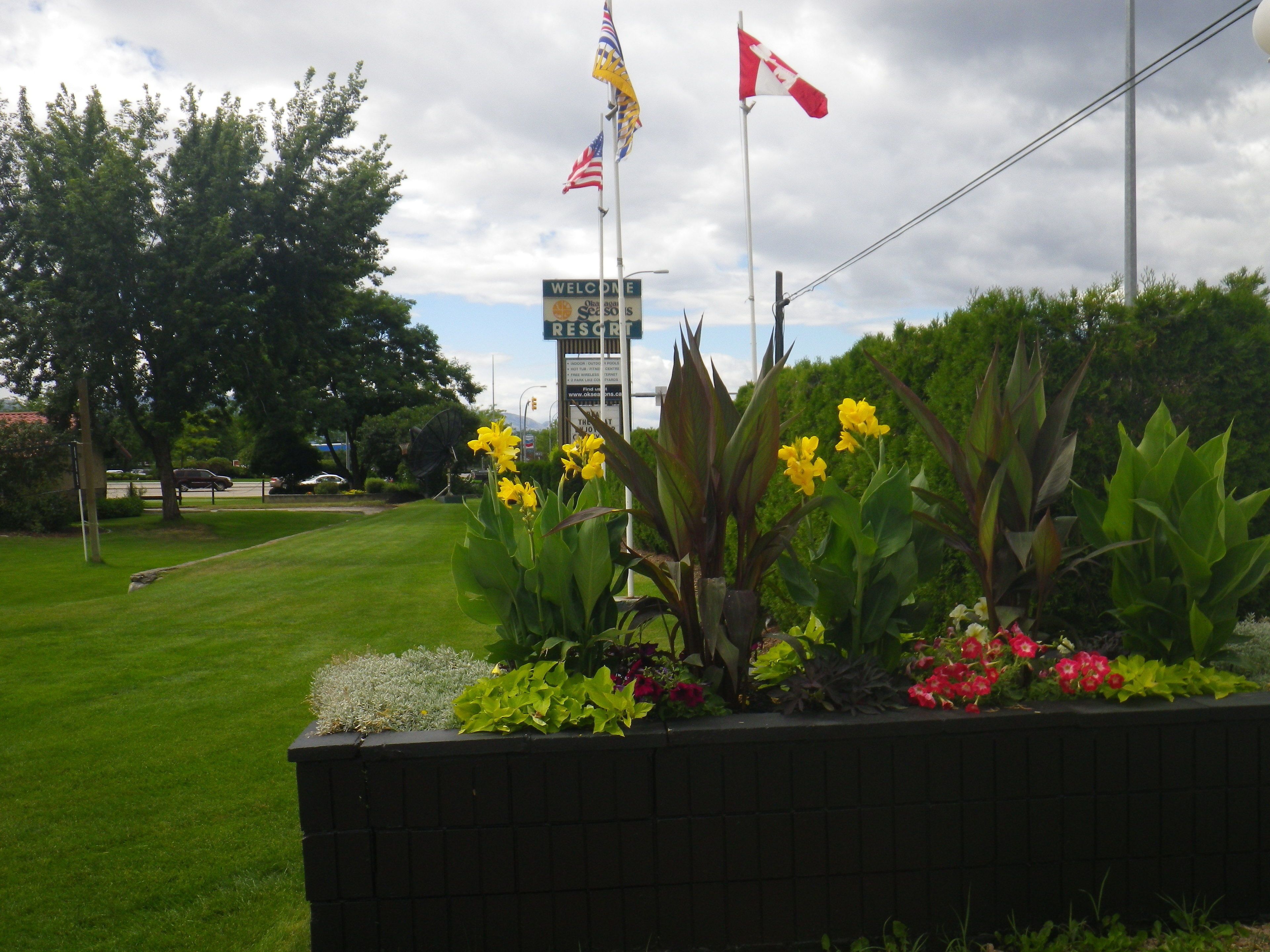 A landscaped area with a sign for a resort, featuring a flagpole with multiple flags, a well-maintained lawn, and various plants.