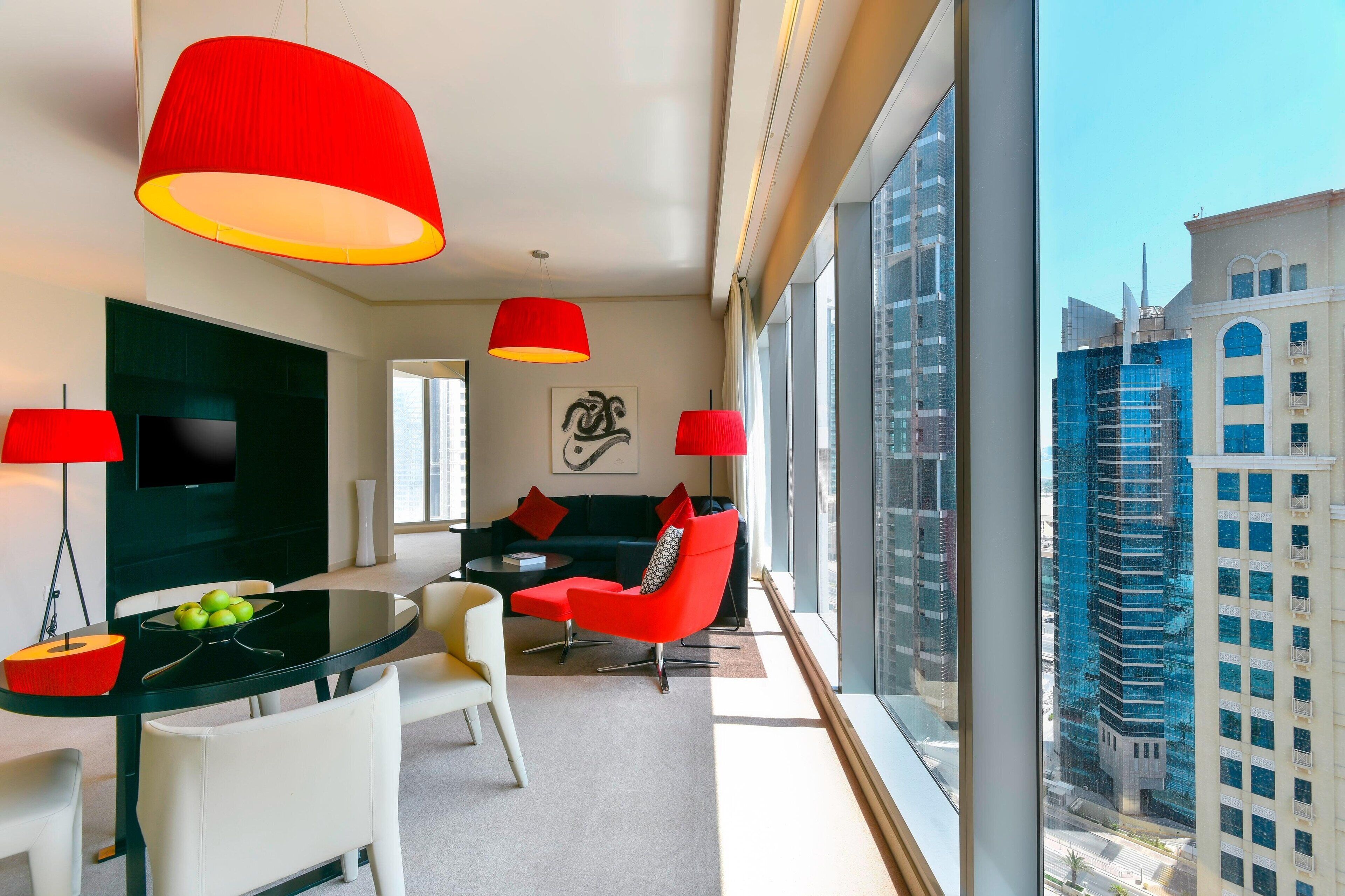 A modern hotel room with a dining area, red lamps, a flat-screen TV, and a city view.