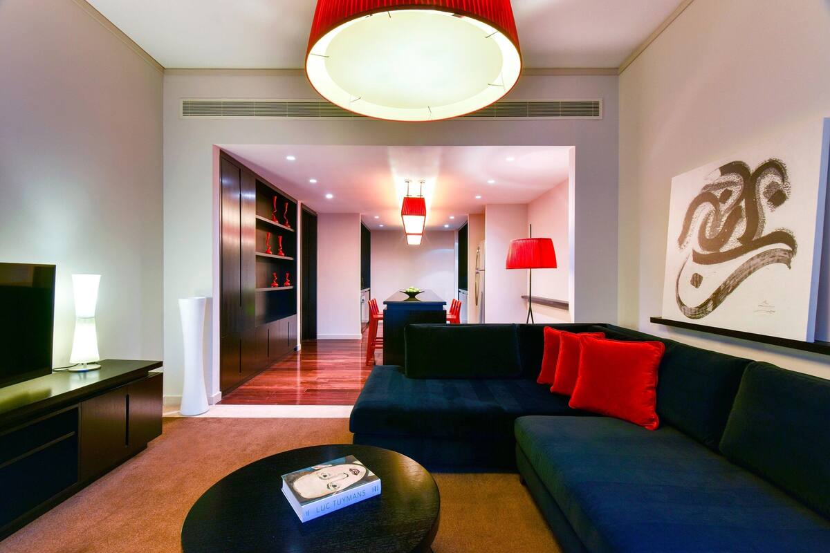 A modern living room with a dark blue sofa, red cushions, a black coffee table with a book, a red floor lamp, and a large abstract painting on the wall.