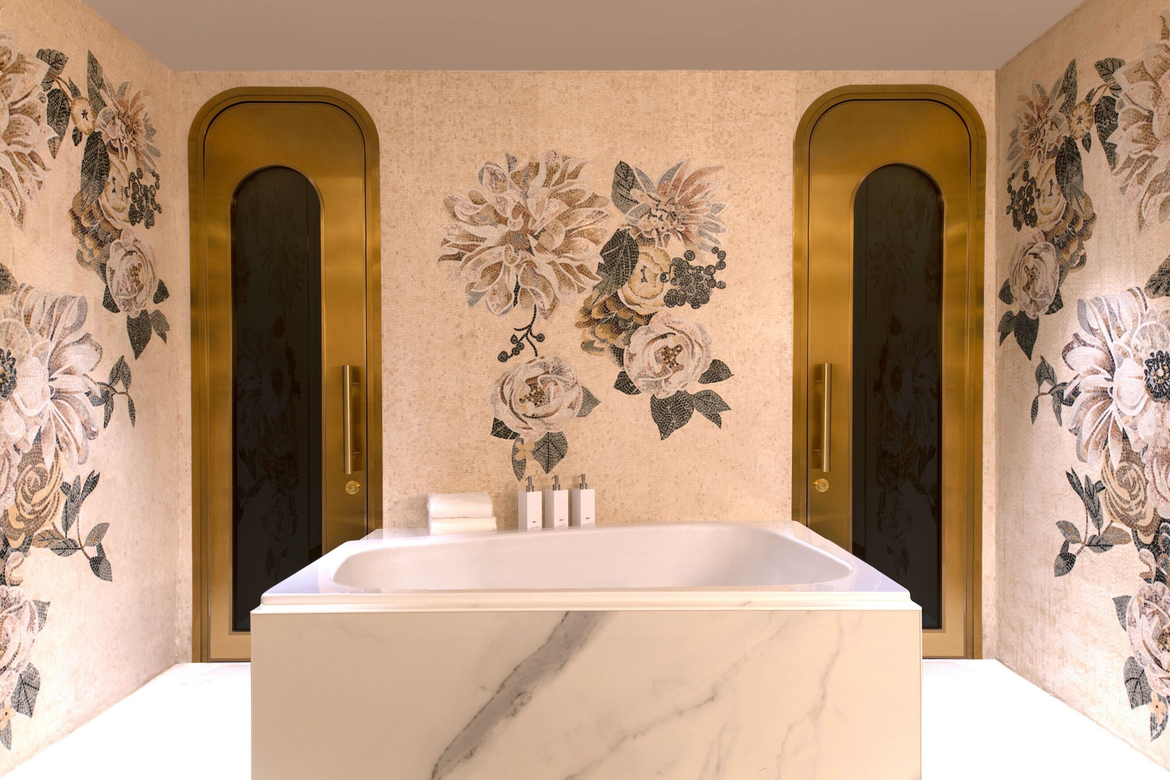 A bathroom with a marble bathtub, floral wall art, and two gold doors.