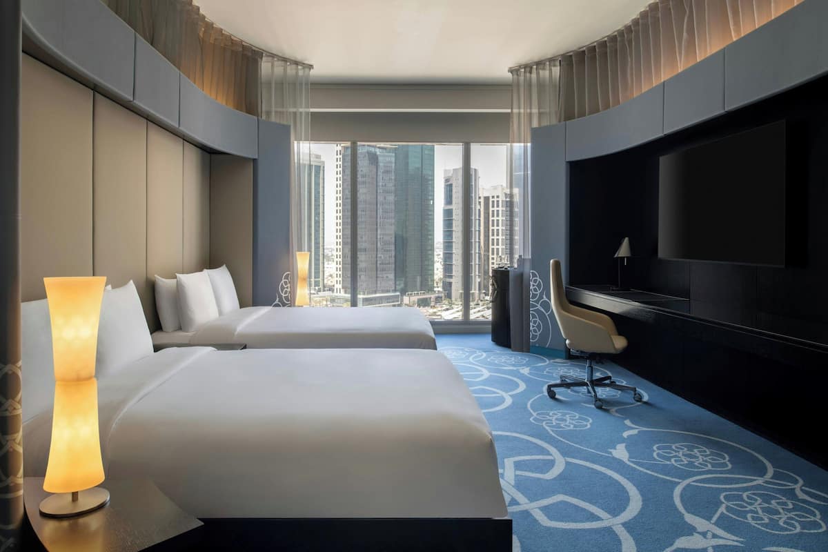 A hotel room with two beds, a TV, a desk, and a view of tall buildings.