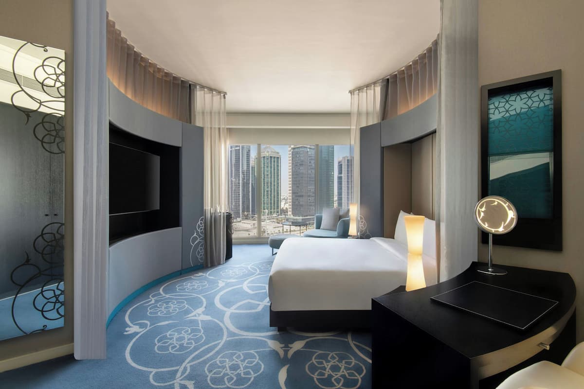 A hotel room with a large bed, a desk with a mirror, a view of the cityscape, and a patterned carpet.