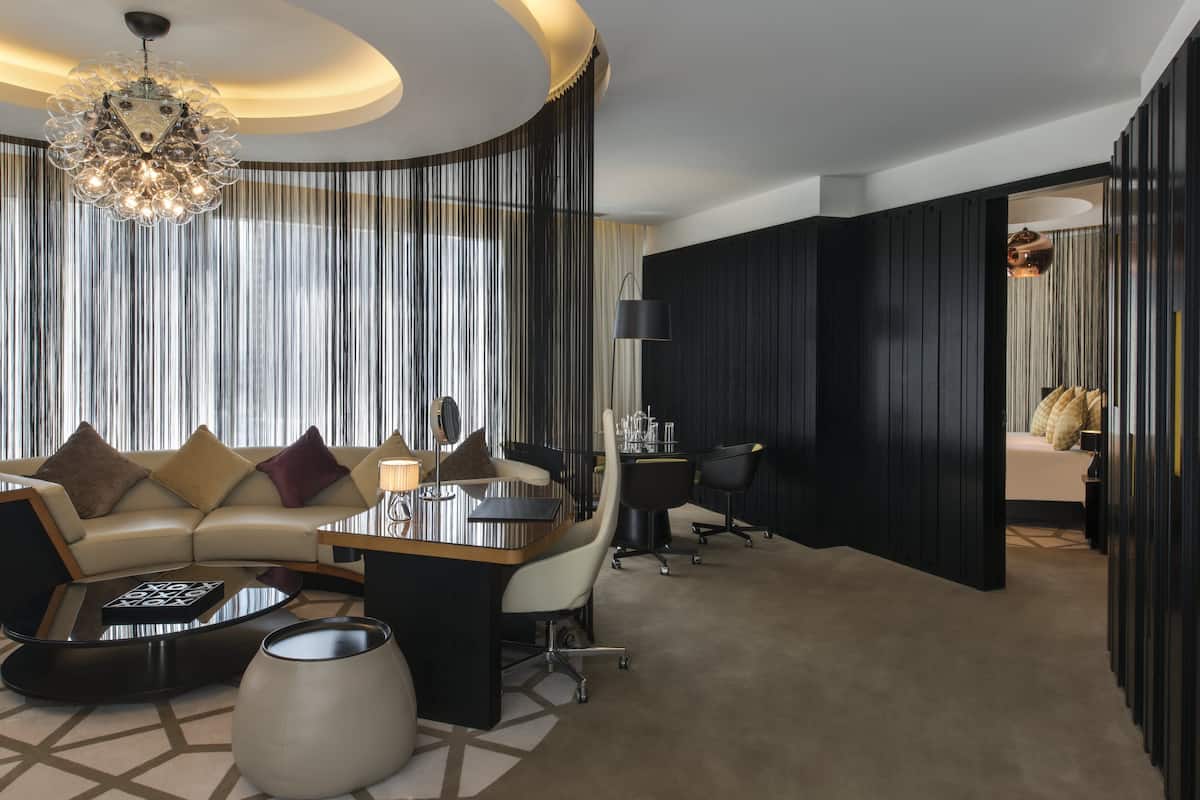 A modern hotel room with a curved ceiling, a chandelier, a sofa set, a dining table, and a separate work area with a chair and desk.