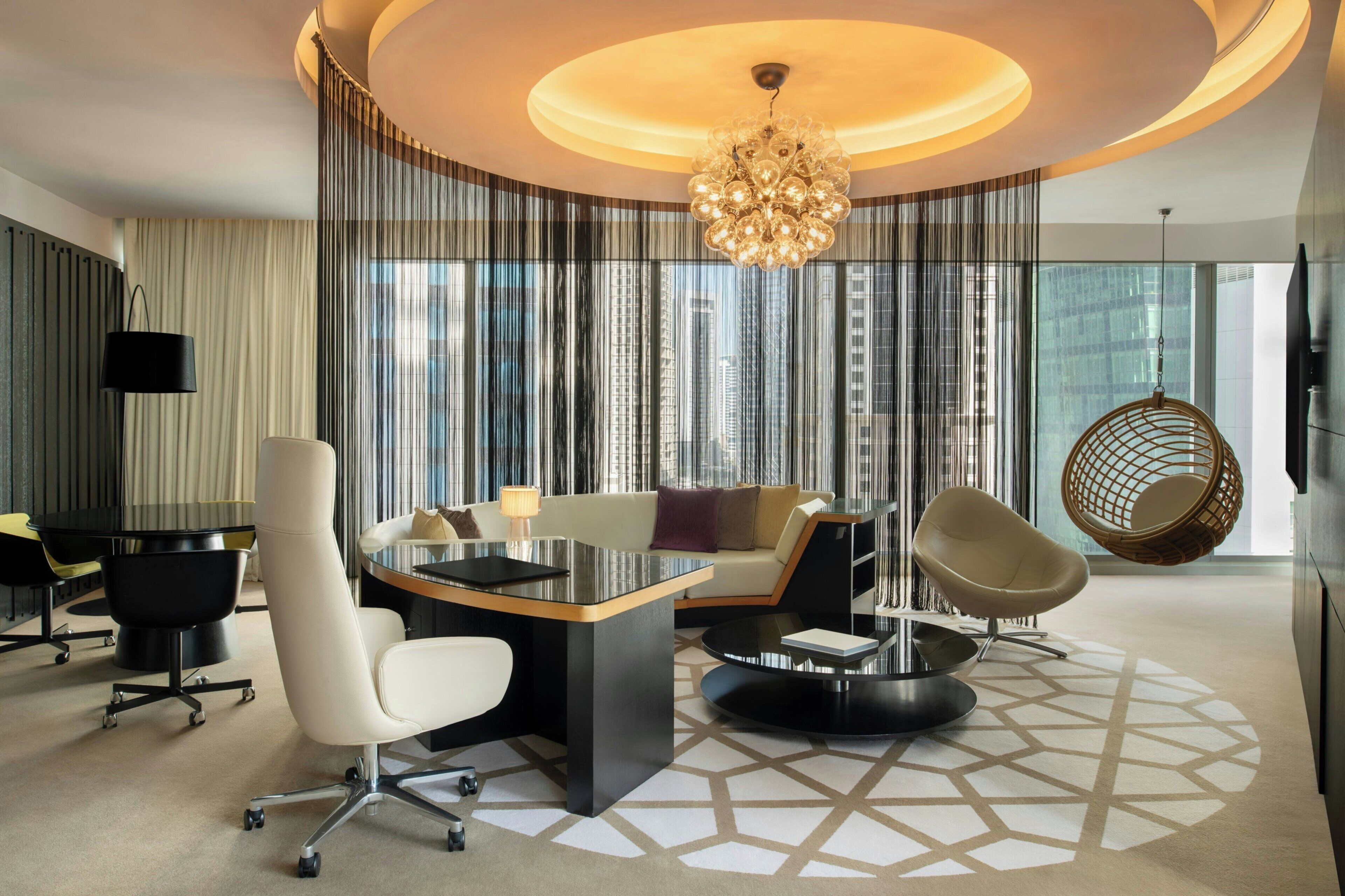 Une chambre d’hôtel moderne avec un bureau rond, un canapé, un fauteuil suspendu et une grande fenêtre donnant sur des immeubles.