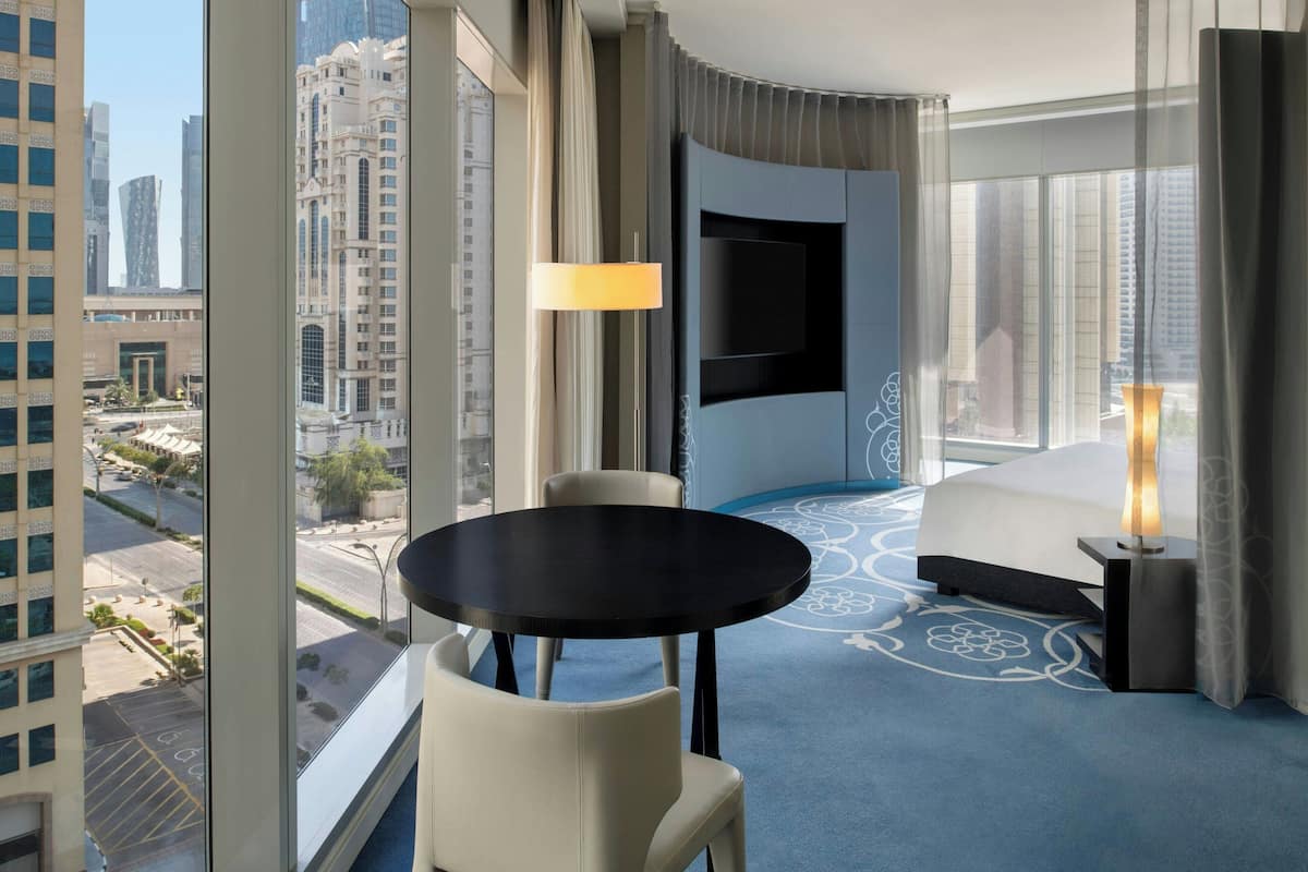 A hotel room with a large window, a round table, a sofa, and a TV.