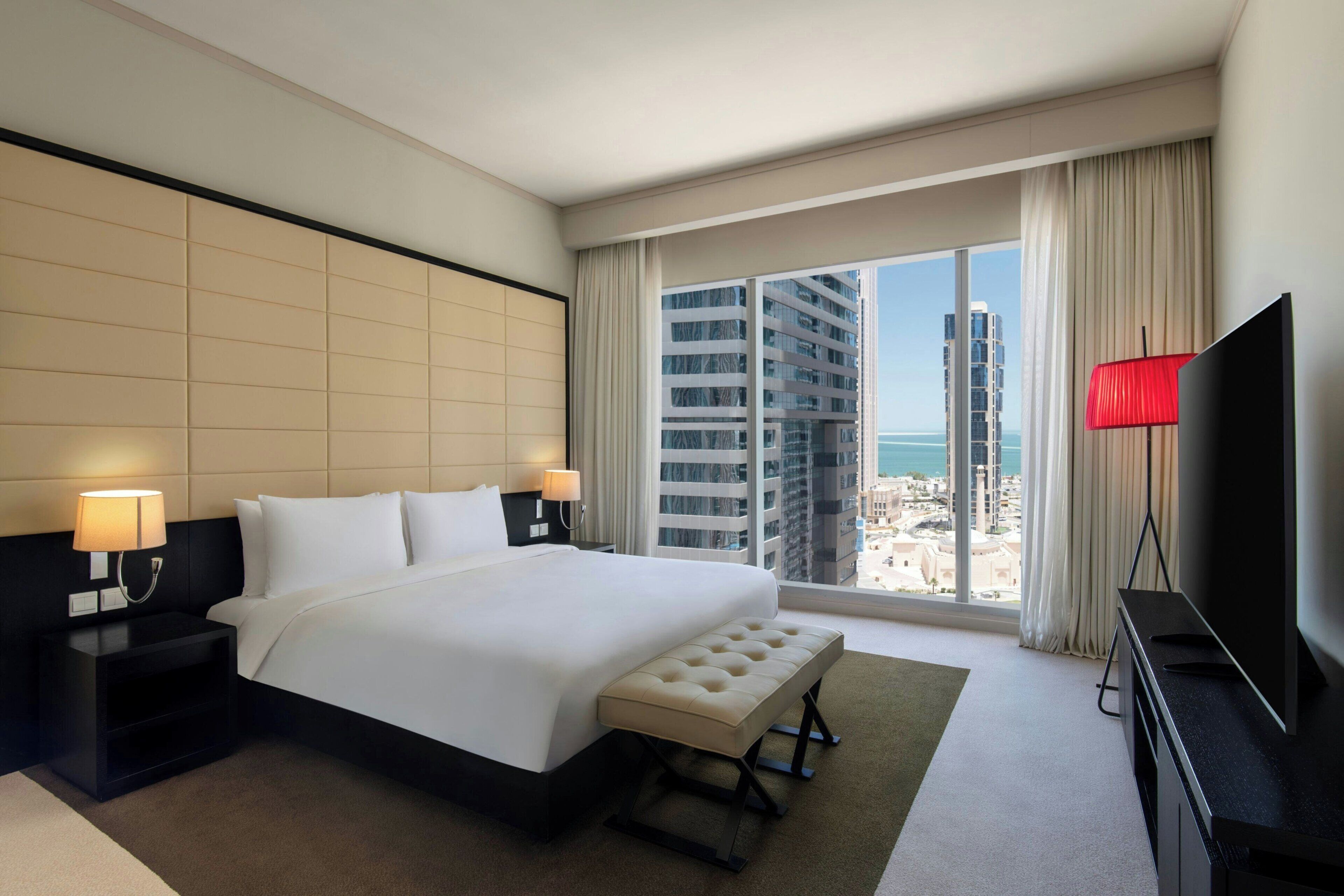 Quarto de hotel com uma cama grande, televisão de tela plana, mesa de cabeceira, abajur e vista para a cidade.