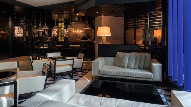 Un salon moderne avec un canapé, des fauteuils et une table basse. On aperçoit une cheminée et une lampe au fond.