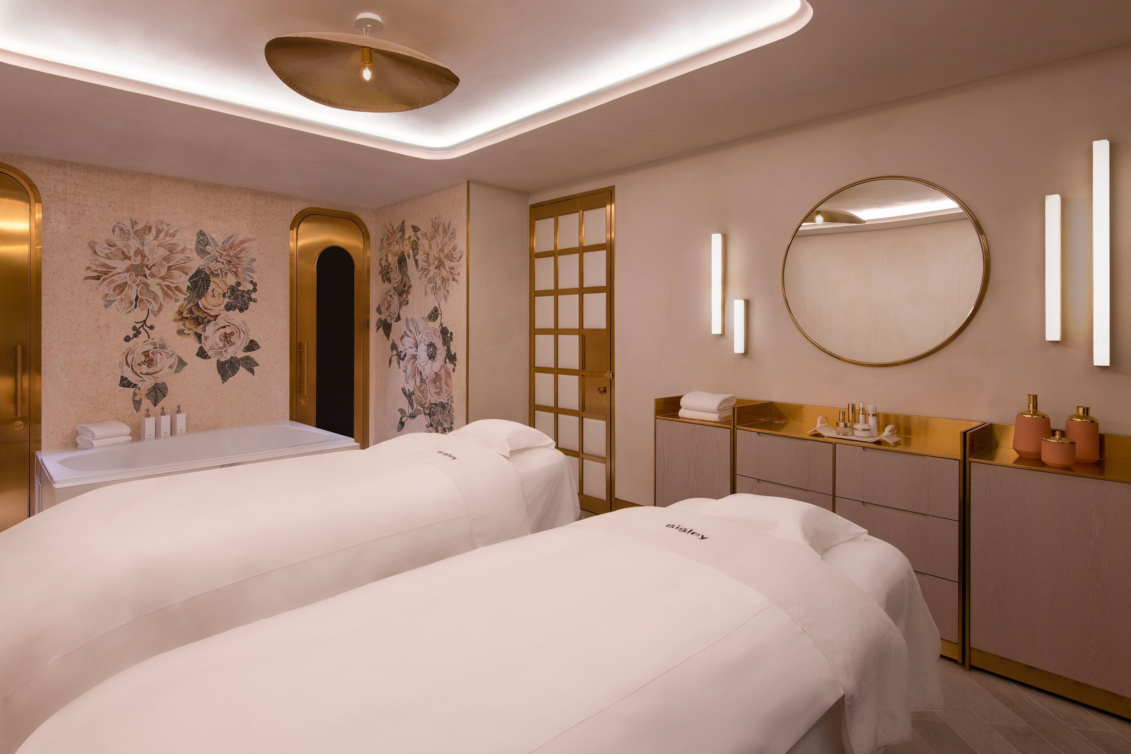 A spa room with two treatment beds, a gold-framed mirror, and a wall-mounted vanity with a sink.