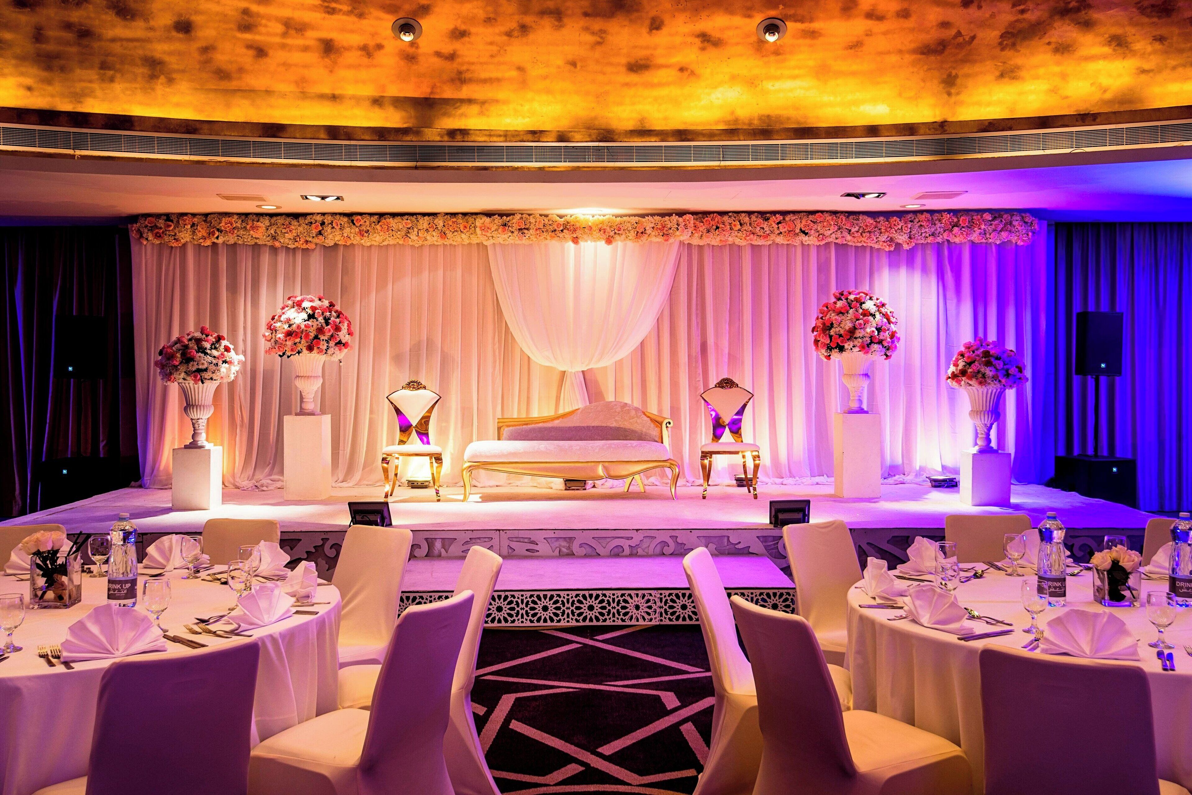 A stage with a bed, floral arrangements, and a draped curtain backdrop. Tables are set with white tablecloths and chairs.