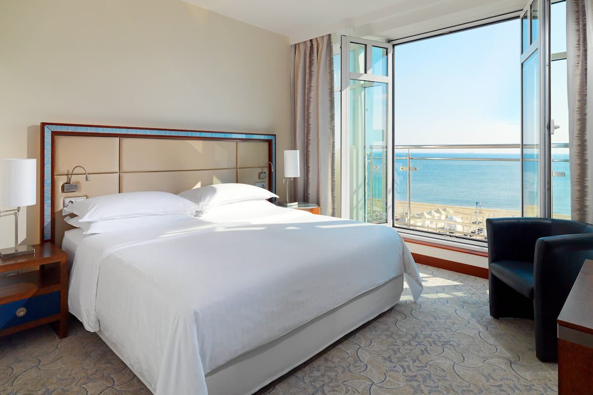 Ein Hotelzimmer mit einem großen Bett, einem Nachttisch, einem Sessel und Blick auf das Meer durch ein großes Fenster.
