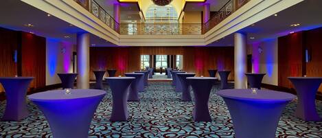 A spacious lobby with a patterned carpet, multiple round tables, and a high ceiling with a decorative light fixture.