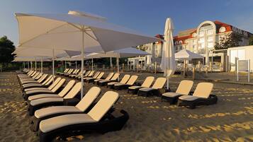 A beach with rows of sun loungers and umbrellas, a hotel building in the background, and a clear sky.