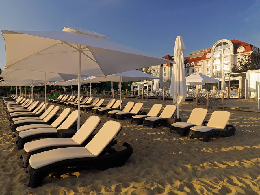 A beach with rows of sun loungers and umbrellas, a hotel building in the background, and a clear sky.